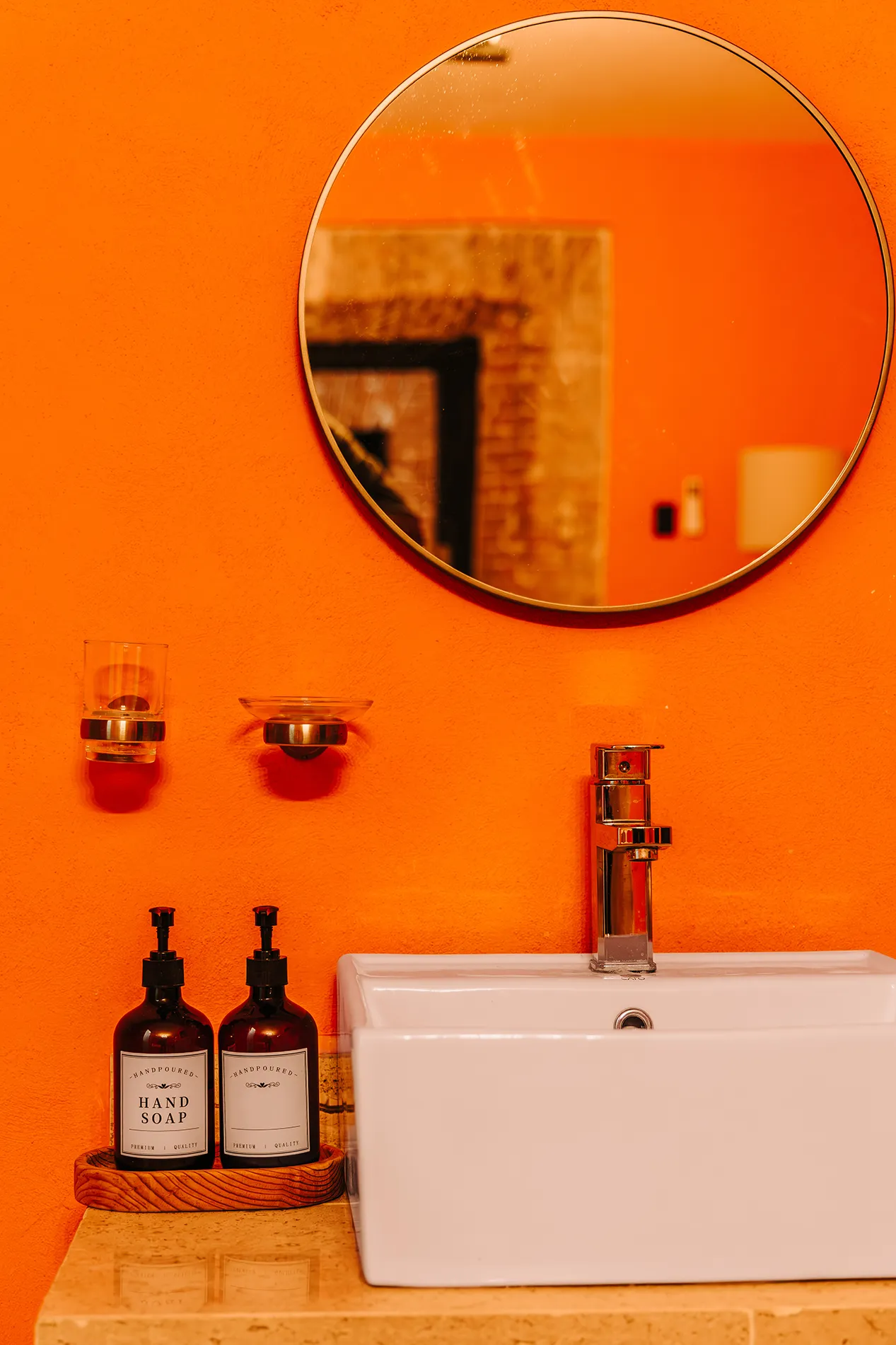 Detalle de lavabo moderno en Casa Grande Rio - Hotel Boutique de Rioverde con espejo circular, dispensadores de jabón artesanal y pared naranja vibrante.