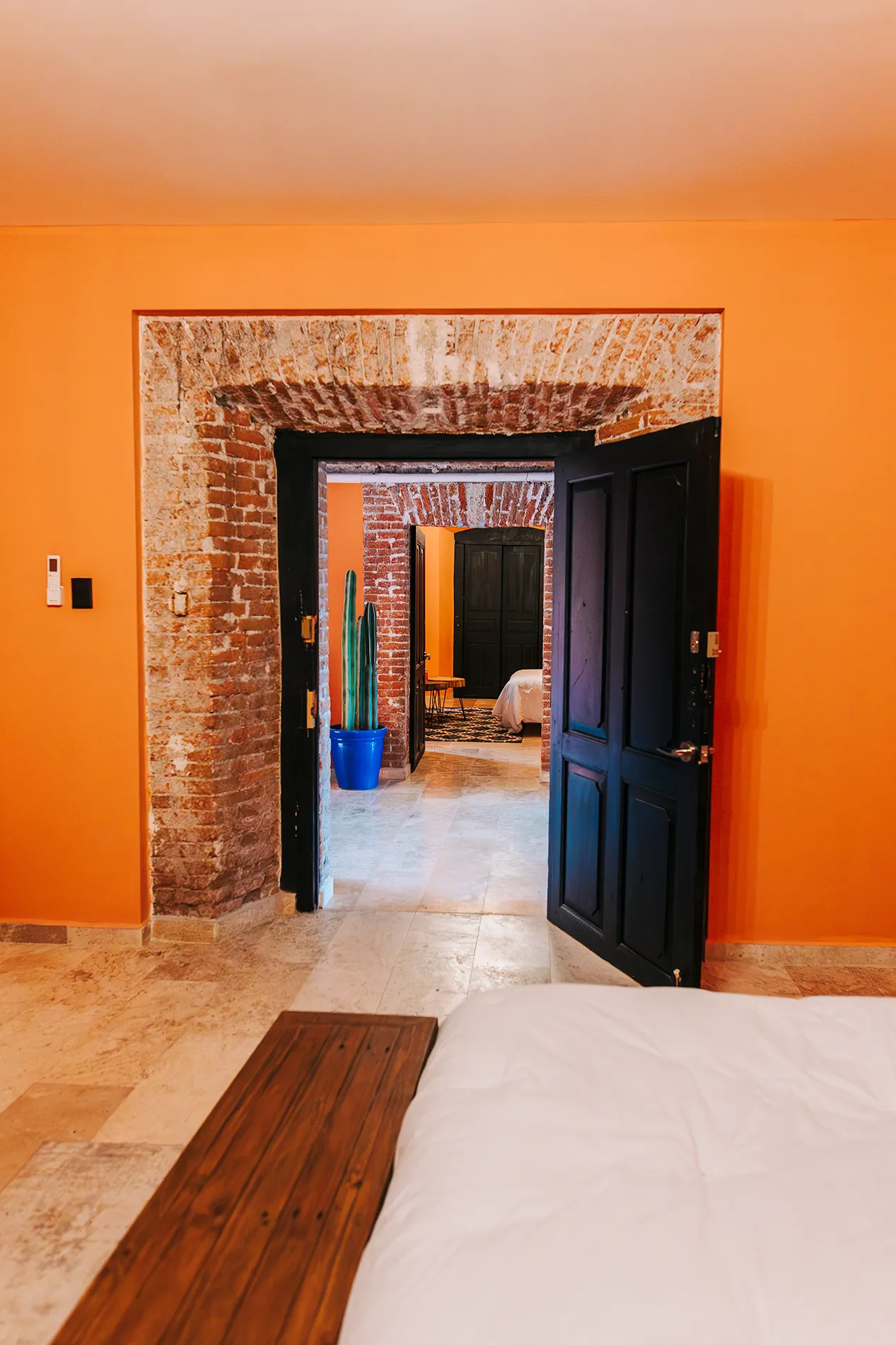 Vista en perspectiva de habitaciones en hotel Casa Grande Rio, Rioverde, a través de arcos de ladrillo rústico, con puertas de madera oscura, paredes naranja y detalles de cactus en macetas azules.