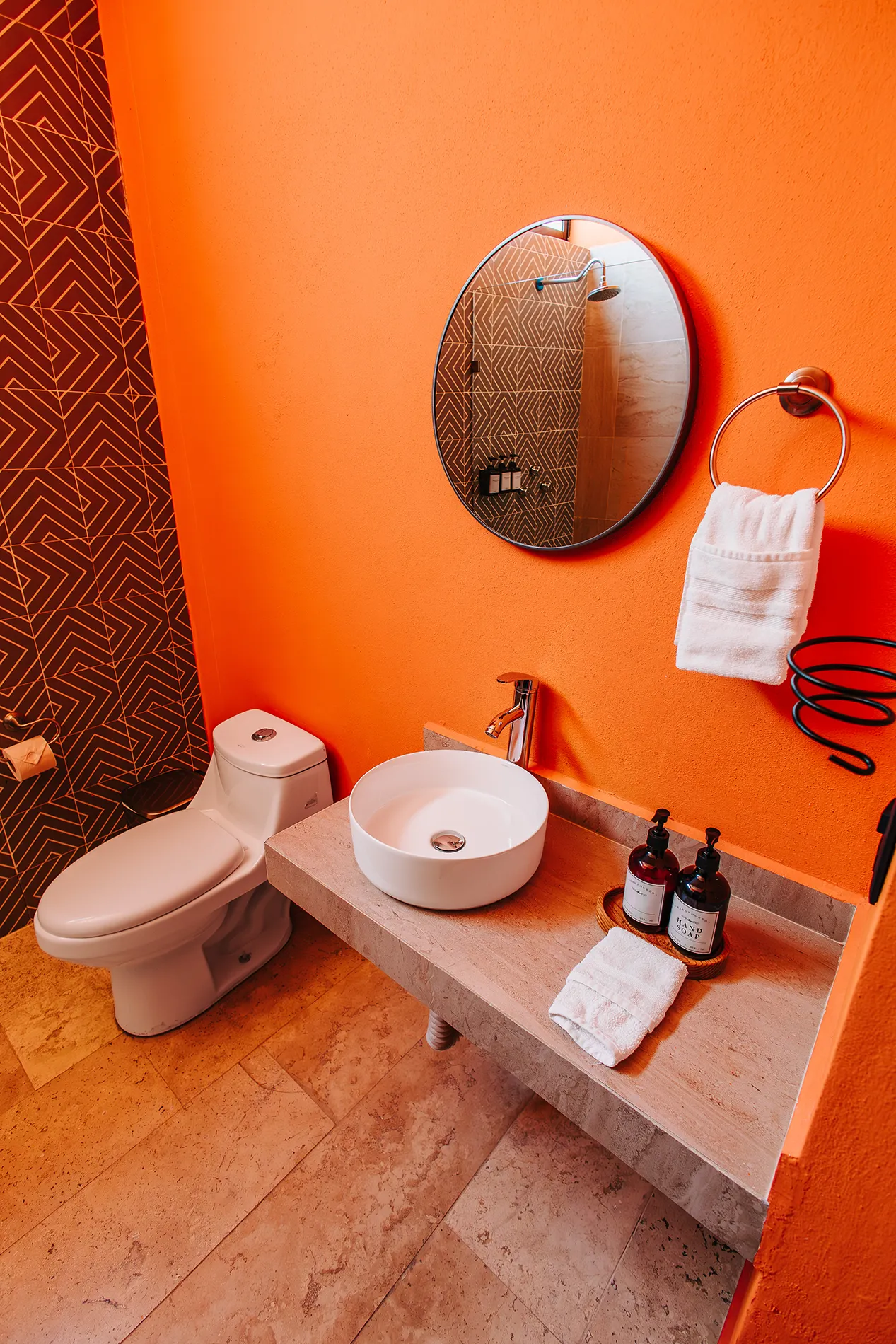 Baño privado y moderno en hotel de Rioverde, SLP, con paredes naranja vibrante, lavabo de sobreponer circular, inodoro de una pieza y espejo redondo. Destaca el papel tapiz geométrico marrón y los amenities de baño.