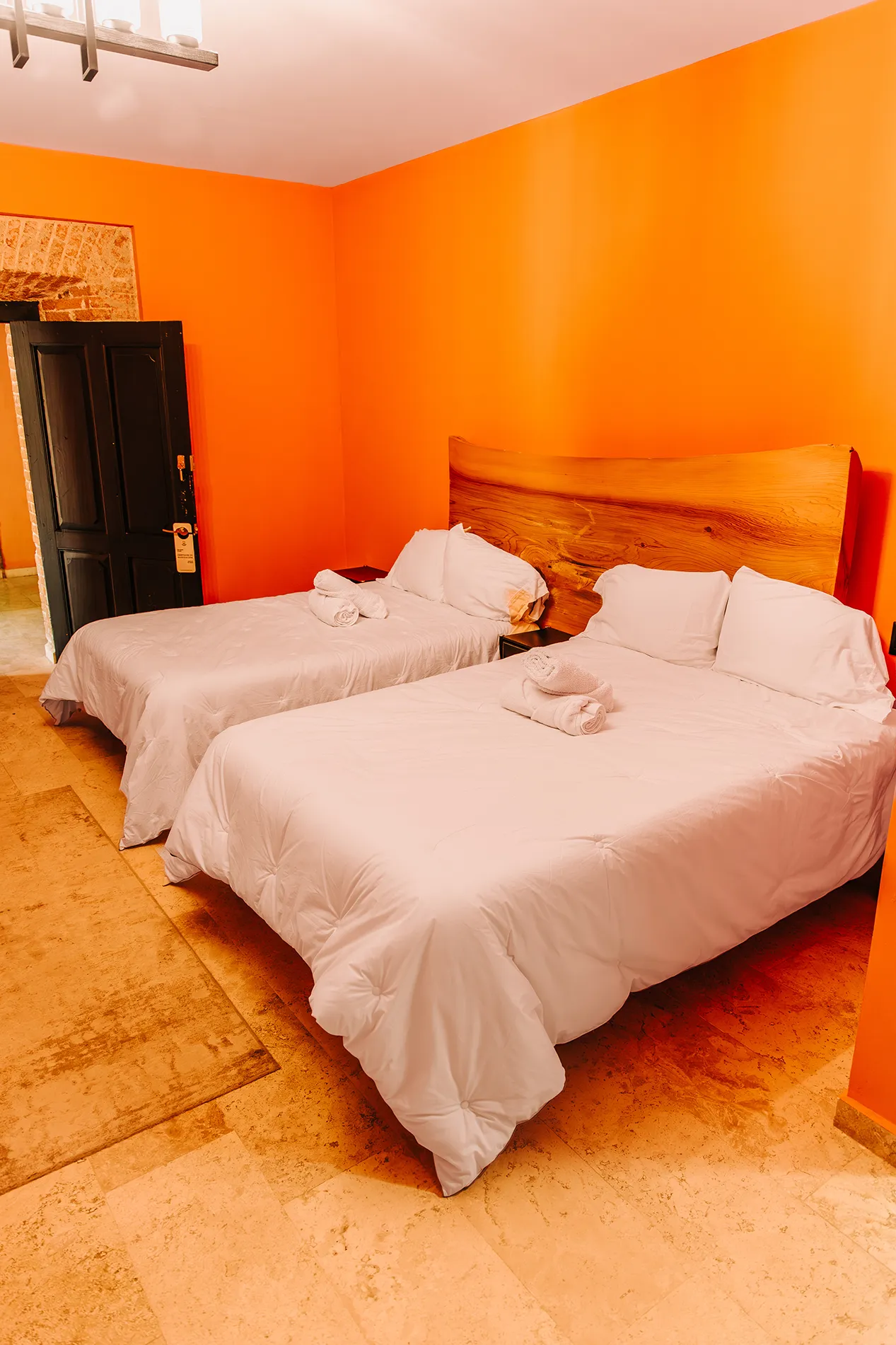 Detalle de habitación 2 con dos camas matrimoniales y cabecera de madera natural de una sola pieza en Casa Grande Rio - Hotel Boutique de Rioverde, SLP, con paredes naranja y piso de mármol travertino.
