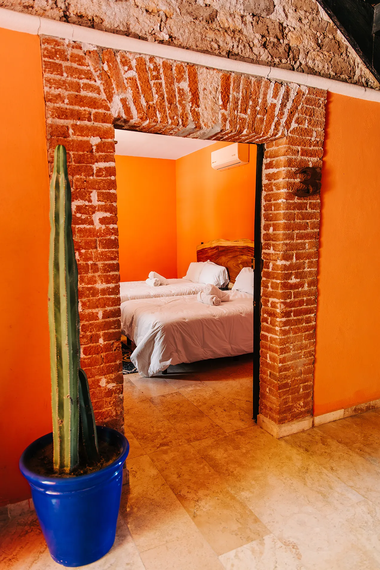Detalle de entrada rústica a habitación 3 de hotel Casa Grande Río en Rioverde, SLP, con arco de ladrillo artesanal, cactus columnar alto en maceta azul cobalto y dos camas matrimoniales de fondo con paredes naranja vibrante.