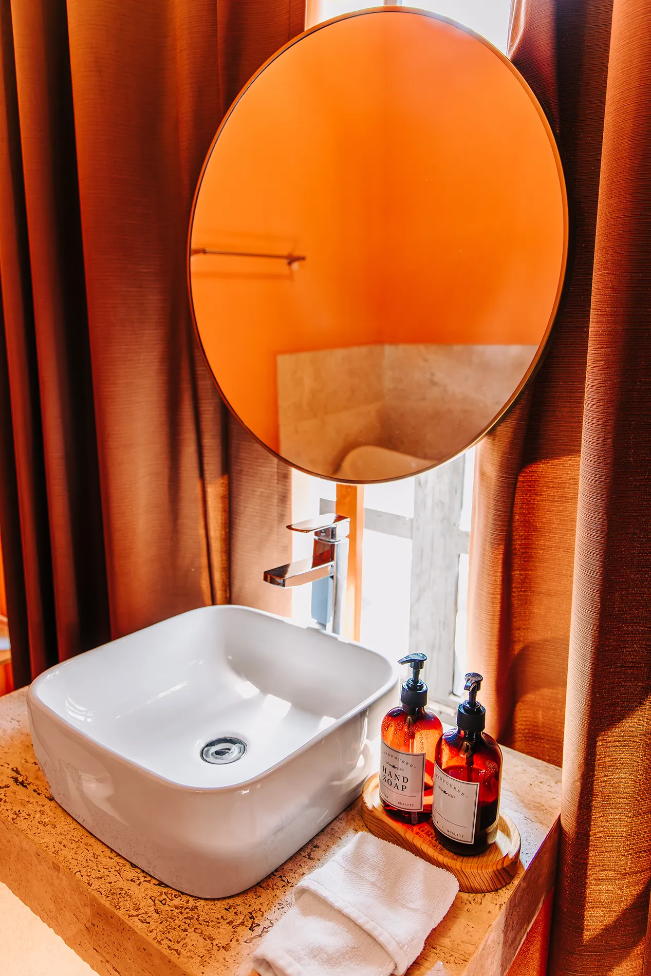 Detalle de baño de hab 8 en Casa Grande Rio - Hotel Boutique de Rioverde con lavabo cuadrado blanco, espejo circular, cortinas terracota y amenidades de jabón premium sobre barra de piedra natural.