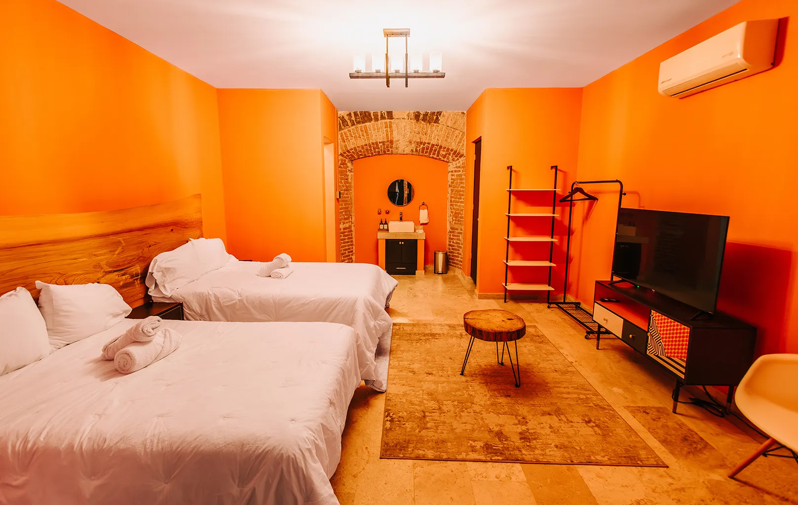 Habitación amplia de Casa Grande Rio, hotel en Rioverde, SLP, con dos camas matrimoniales, cabecera de madera natural, paredes color naranja, área de lavabo bajo arco de ladrillo rústico y Smart TV.