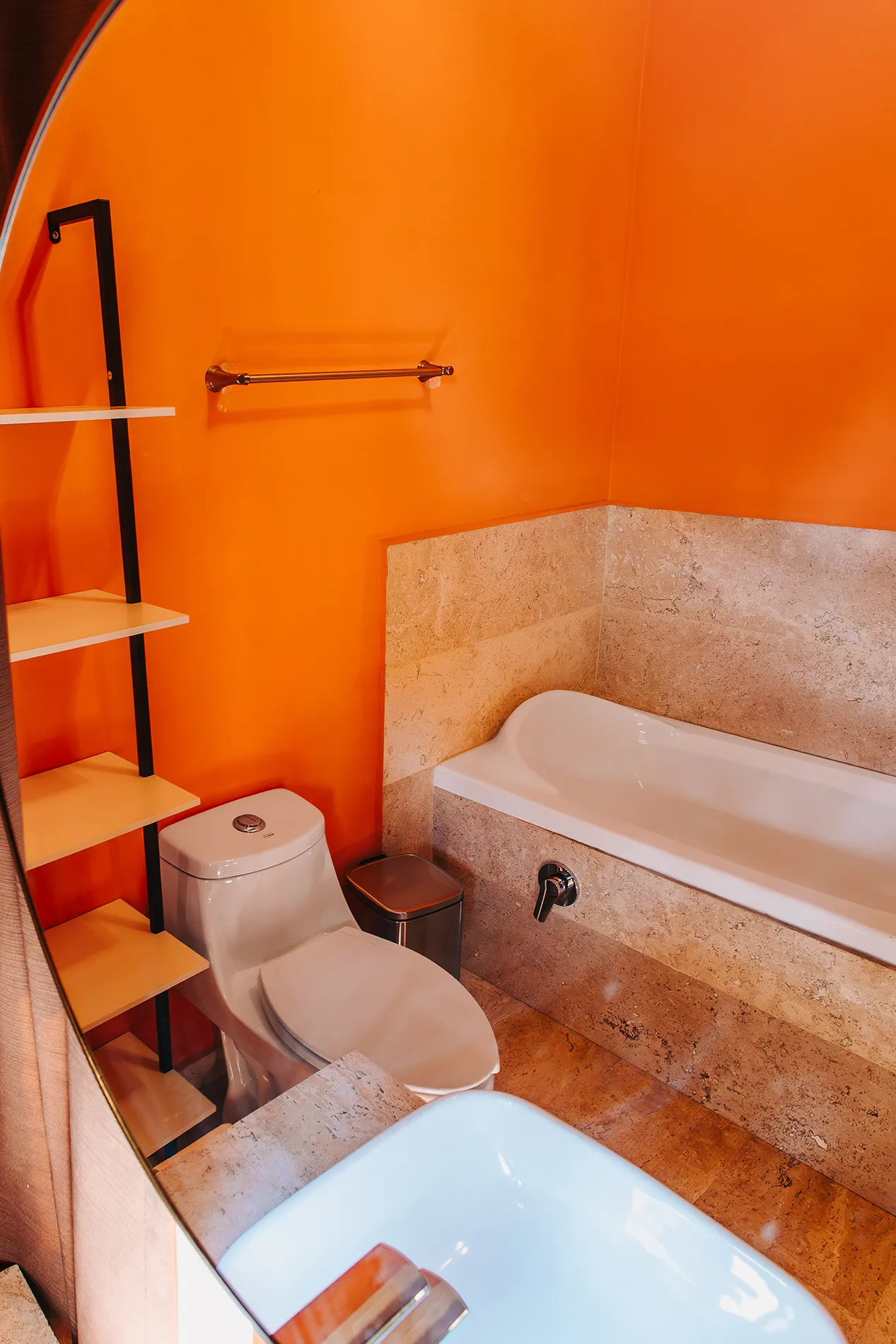 Baño con tina de hidromasaje y acabados en mármol en hotel boutique de Rioverde, SLP, con paredes naranja vibrante, estantería minimalista y sanitarios modernos.